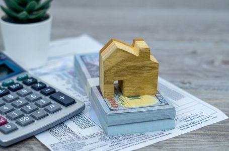 Wooden block house sitting atop stack of US dollars with paperwork and calculator in background